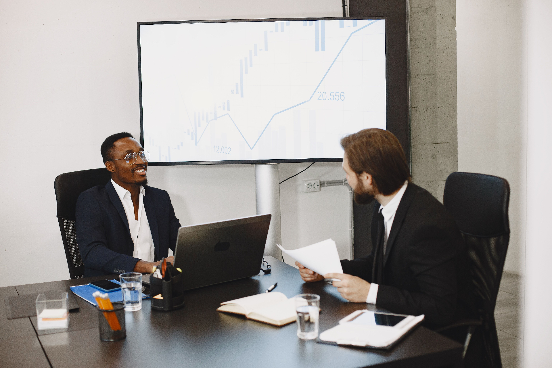 Two investors in a boardroom reviewing startup financials on a laptop and printed reports, with a market growth chart trending upward on the screen behind them.