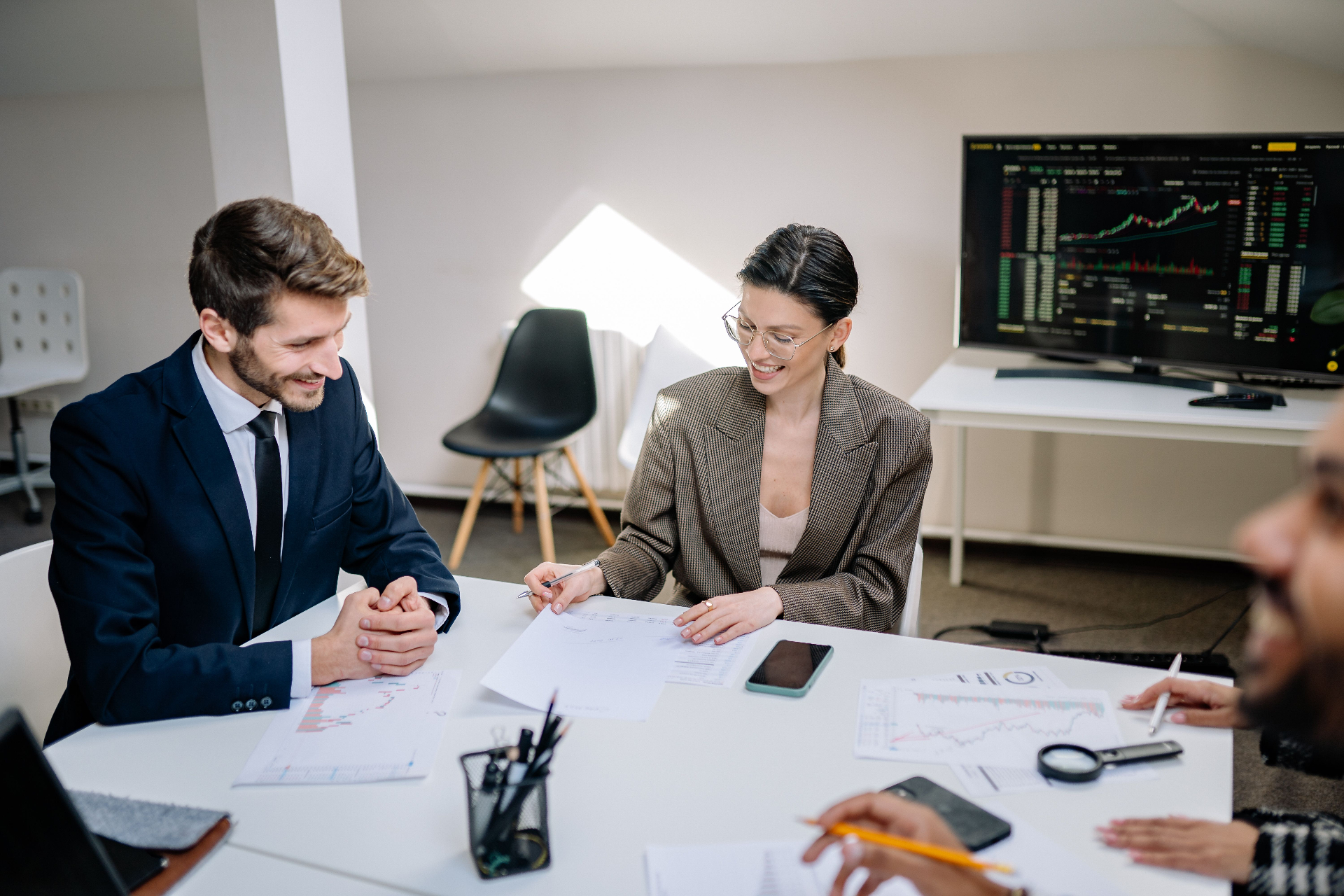 Two early-stage investors reviewing a founder's financial documents and charts across a meeting room table, with a market data screen in the background.